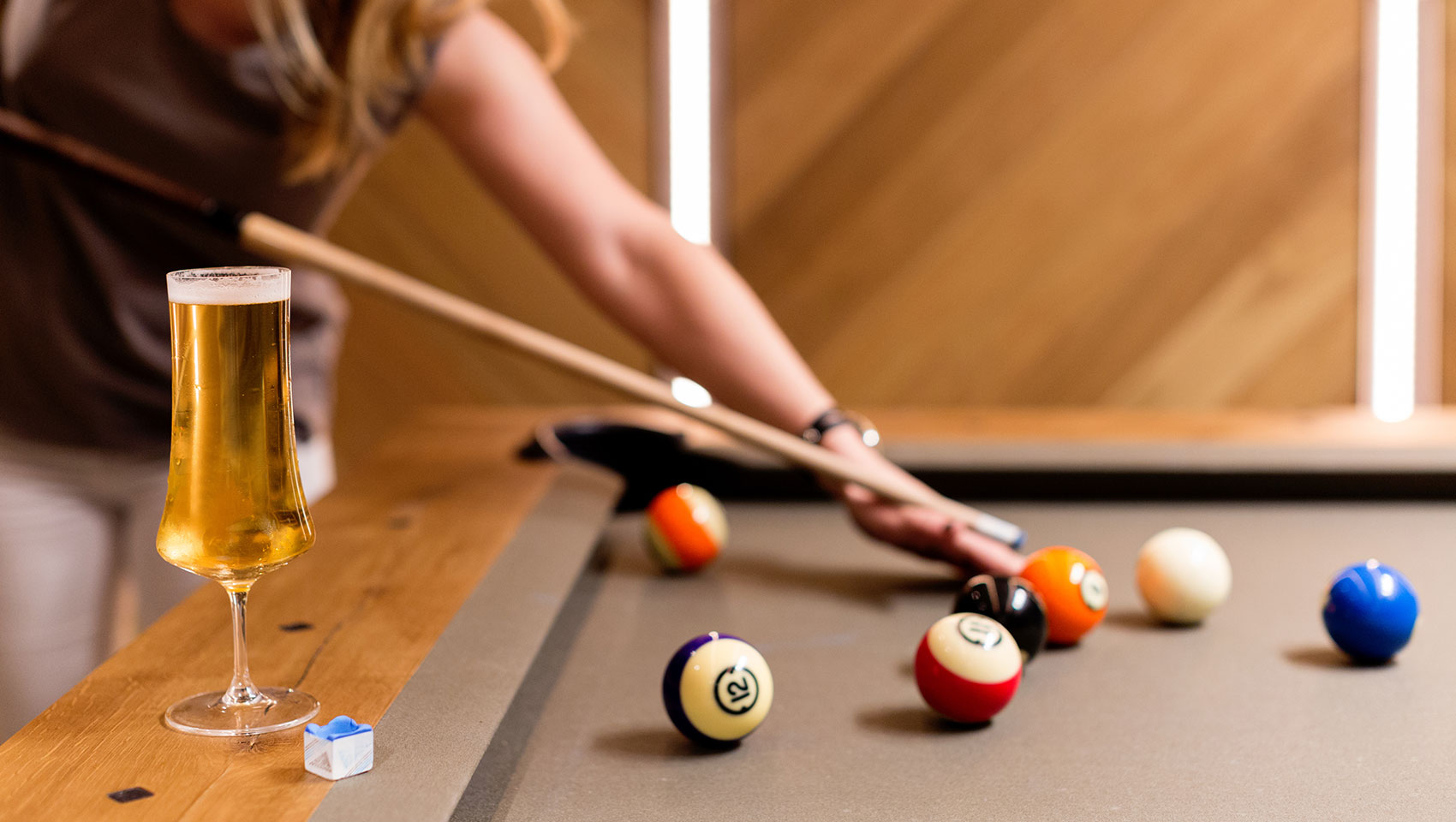 Close up of cocktail glass and pool cue chalk, a woman plays pool out of focus in the background