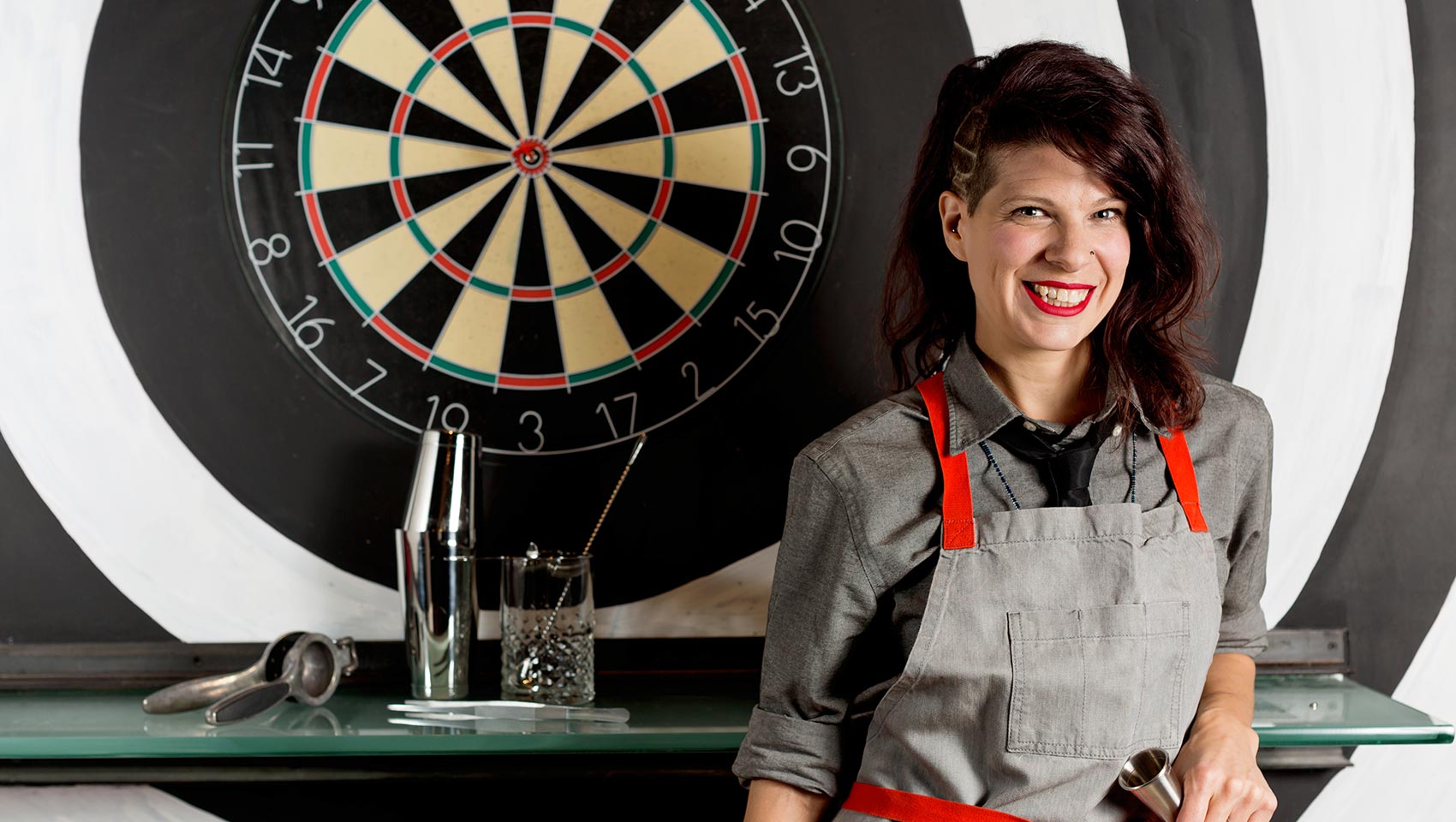 Female bartender, smiling, stands in front of a dart board